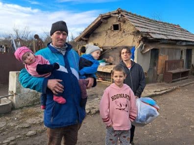 Un tată cu trei copii în brațe, în fața casei familiei, ținând un pachet primit de la caravană